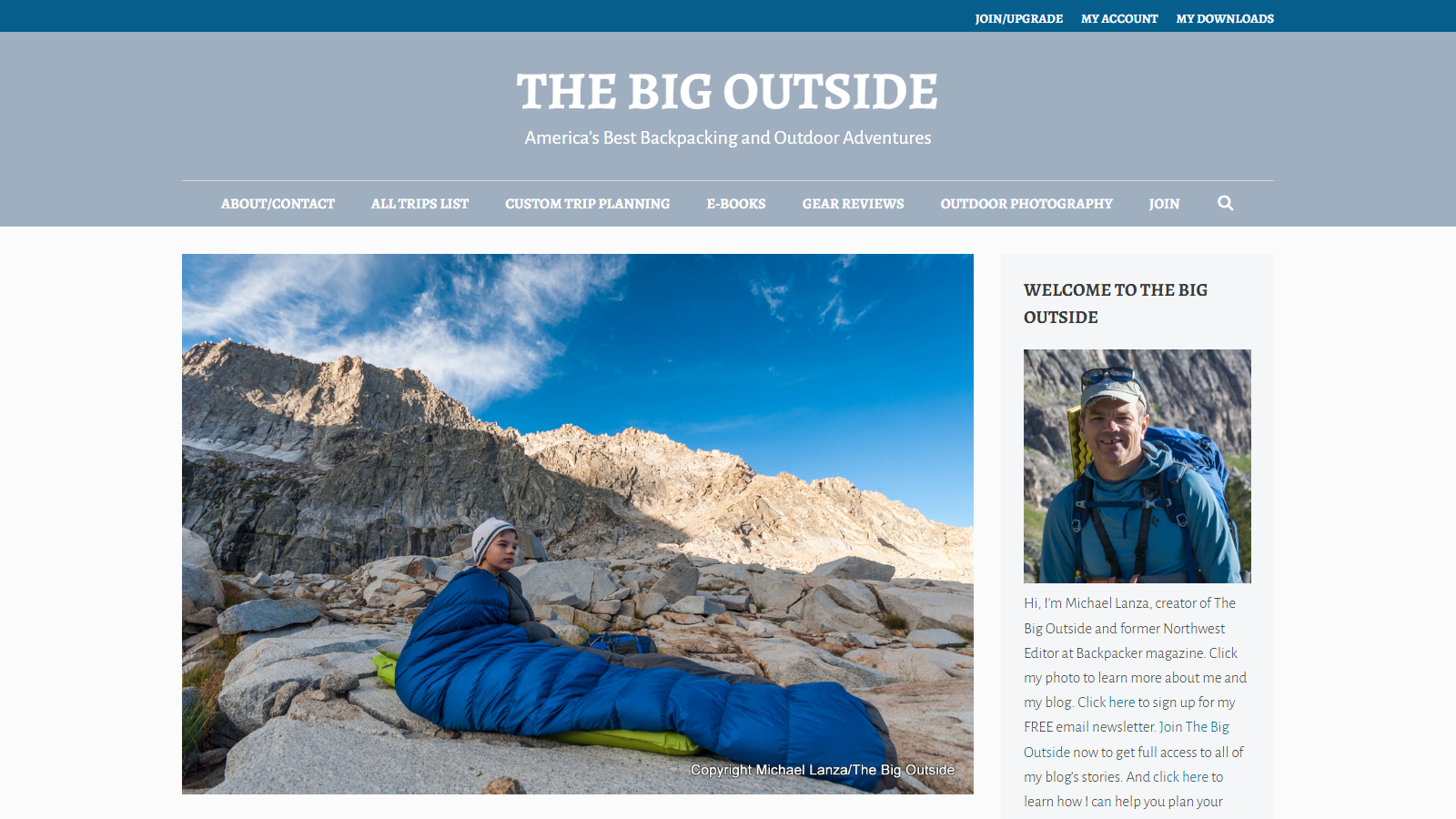 The Big Outside's blue-and-grey masthead above a photo of a young hiker in a grey beanie sitting zipped into a blue down sleeping bag on a green sleeping pad among granite boulders below a sunlit rocky mountain ridge; a sidebar beside it shows a portrait of the blog's author, Michael Lanza, wearing a baseball cap and backpack